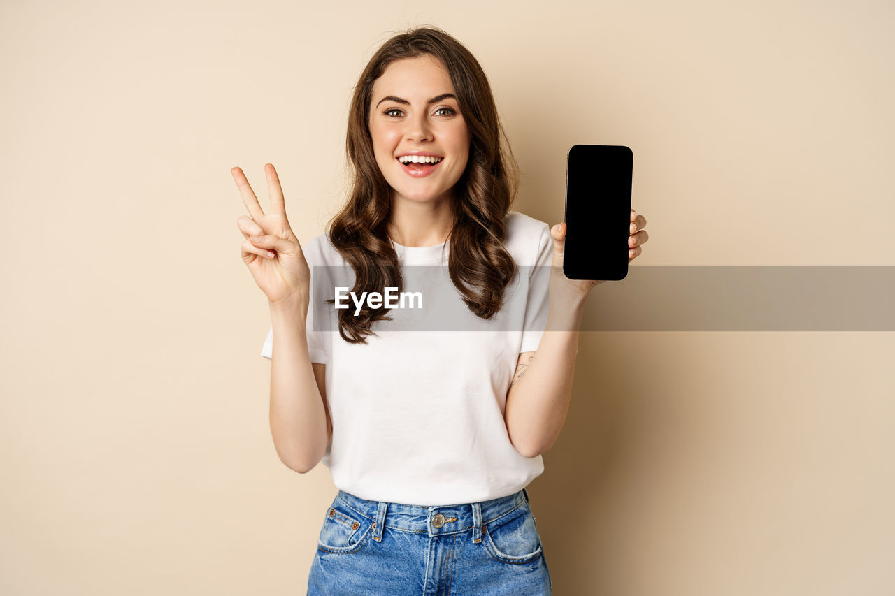 women, one person, portrait, adult, technology, wireless technology, smiling, smartphone, casual clothing, indoors, studio shot, communication, happiness, young adult, portable information device, emotion, mobile phone, photo shoot, jeans, long hair, cheerful, front view, looking at camera, finger, standing, hand, hairstyle, positive emotion, copy space, female, teenager, person, lifestyles, holding, brown hair, clothing, selfie, fun, relaxation, colored background, human face, enjoyment, fashion, joy, arm, waist up, cut out, three quarter length, facial expression, looking