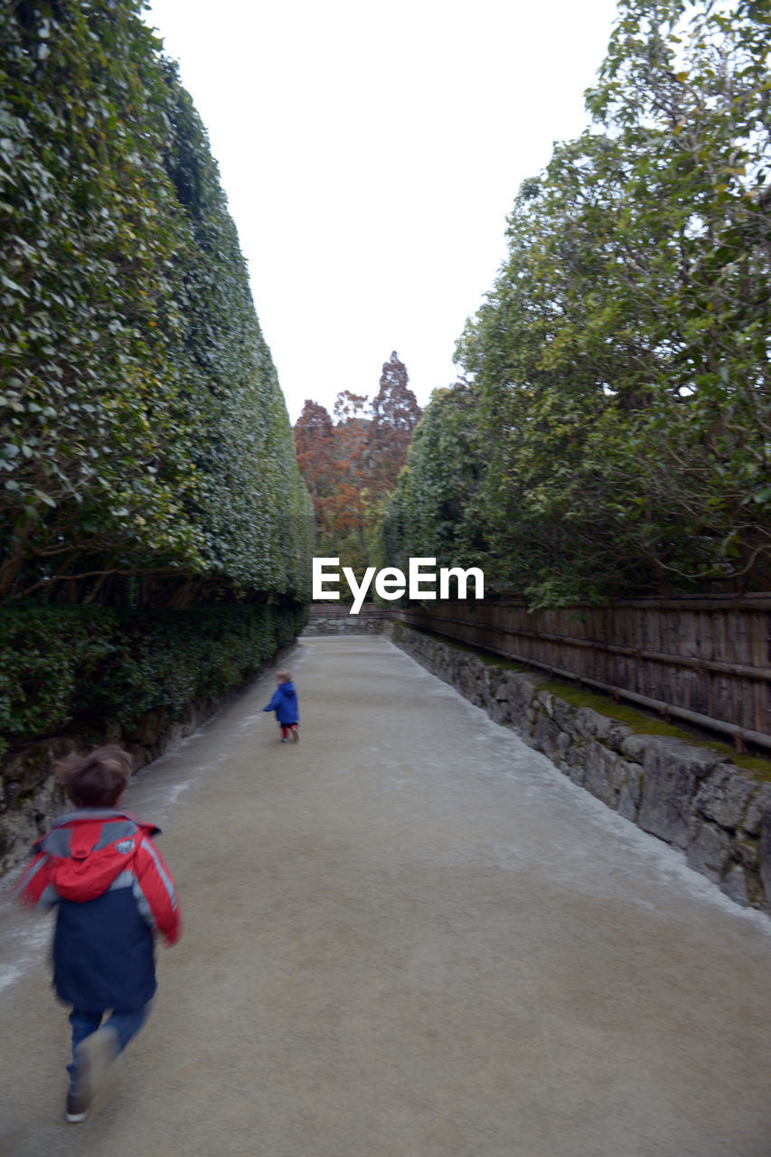 REAR VIEW OF MEN WALKING ON FOOTPATH BY TREES AGAINST SKY