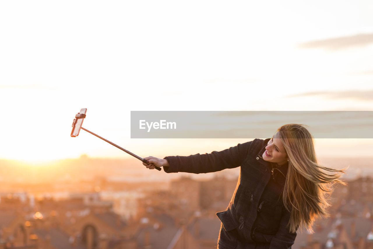 Young woman taking selfie on mobile phone with monopod while standing against sky
