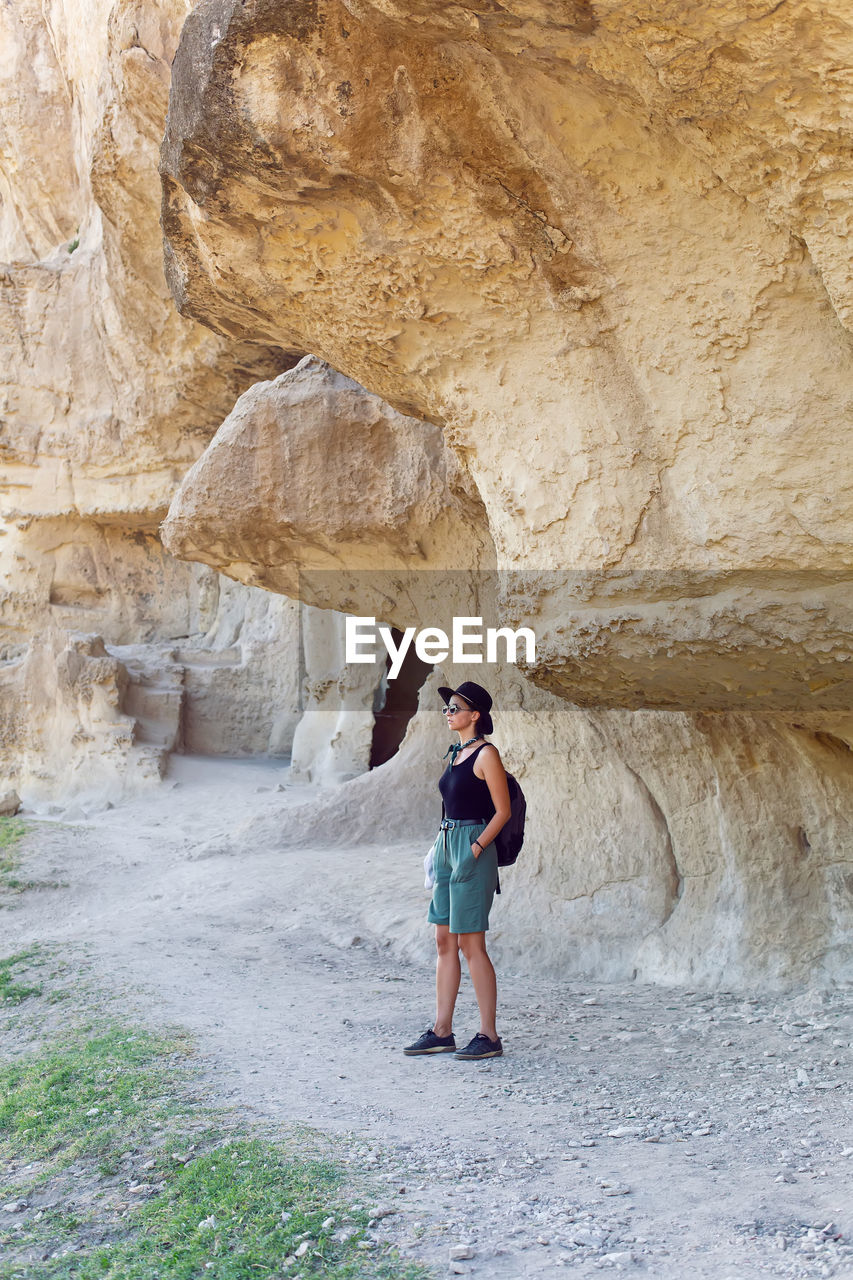 Woman traveler with a backpack and a hat stands next to a rock in the crimea in the summer