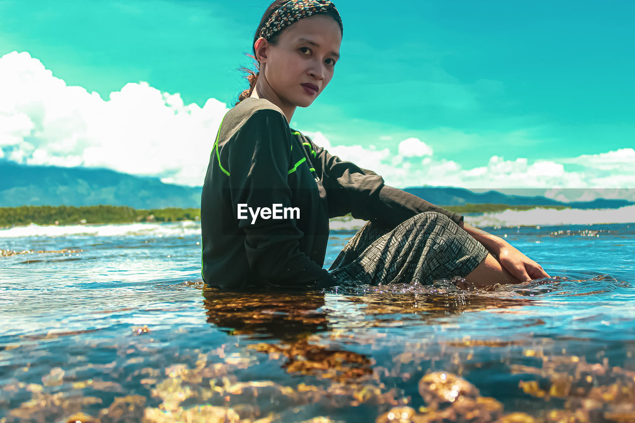 WOMAN SITTING IN SEA AGAINST SKY