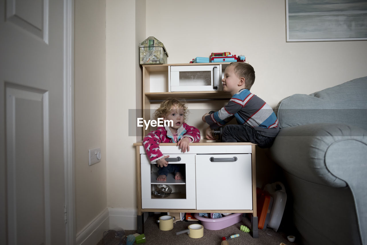 Siblings playing on cabinet by sofa at home