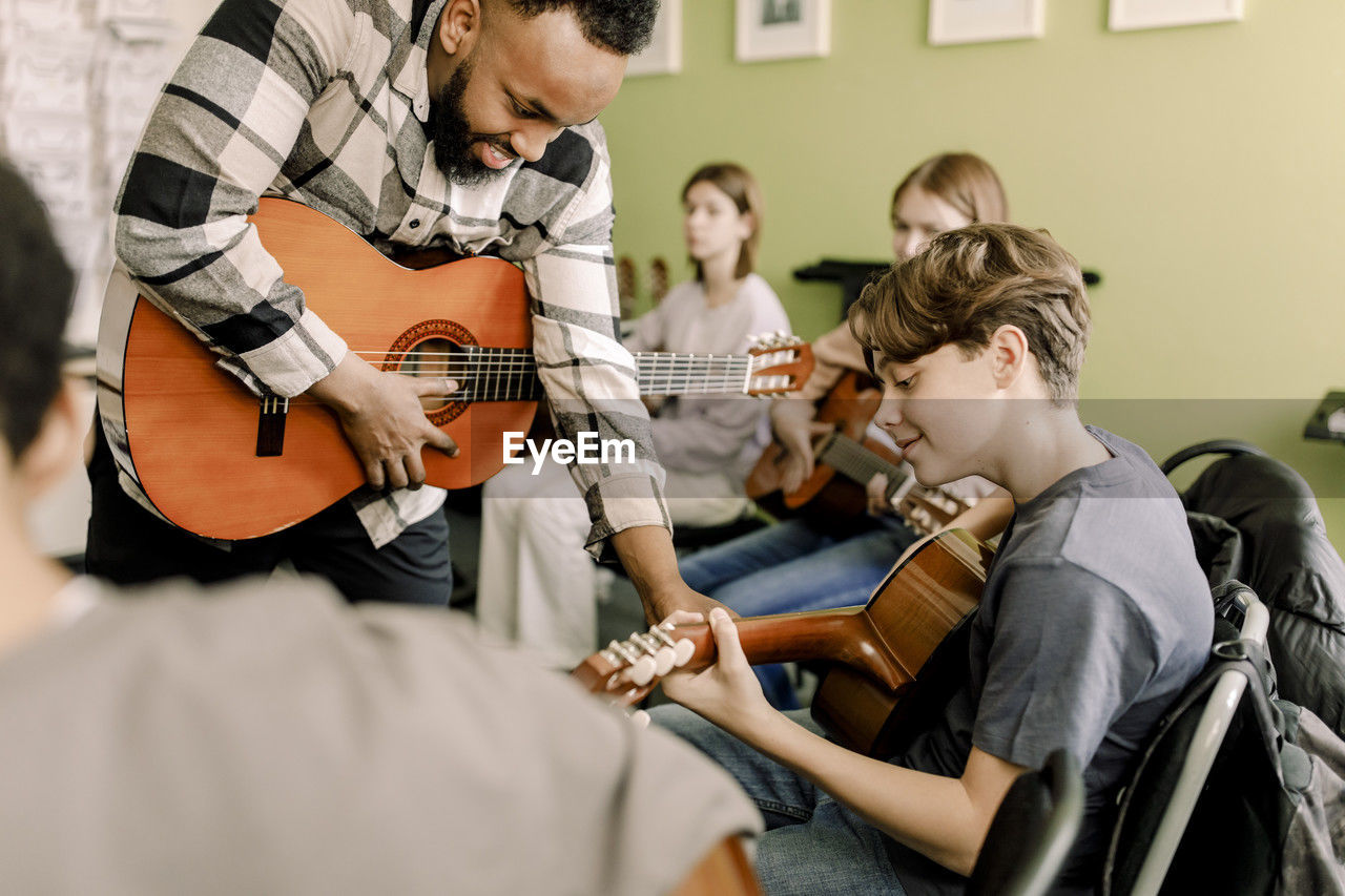 Young male teacher assisting teenage boy playing guitar in music class at high school