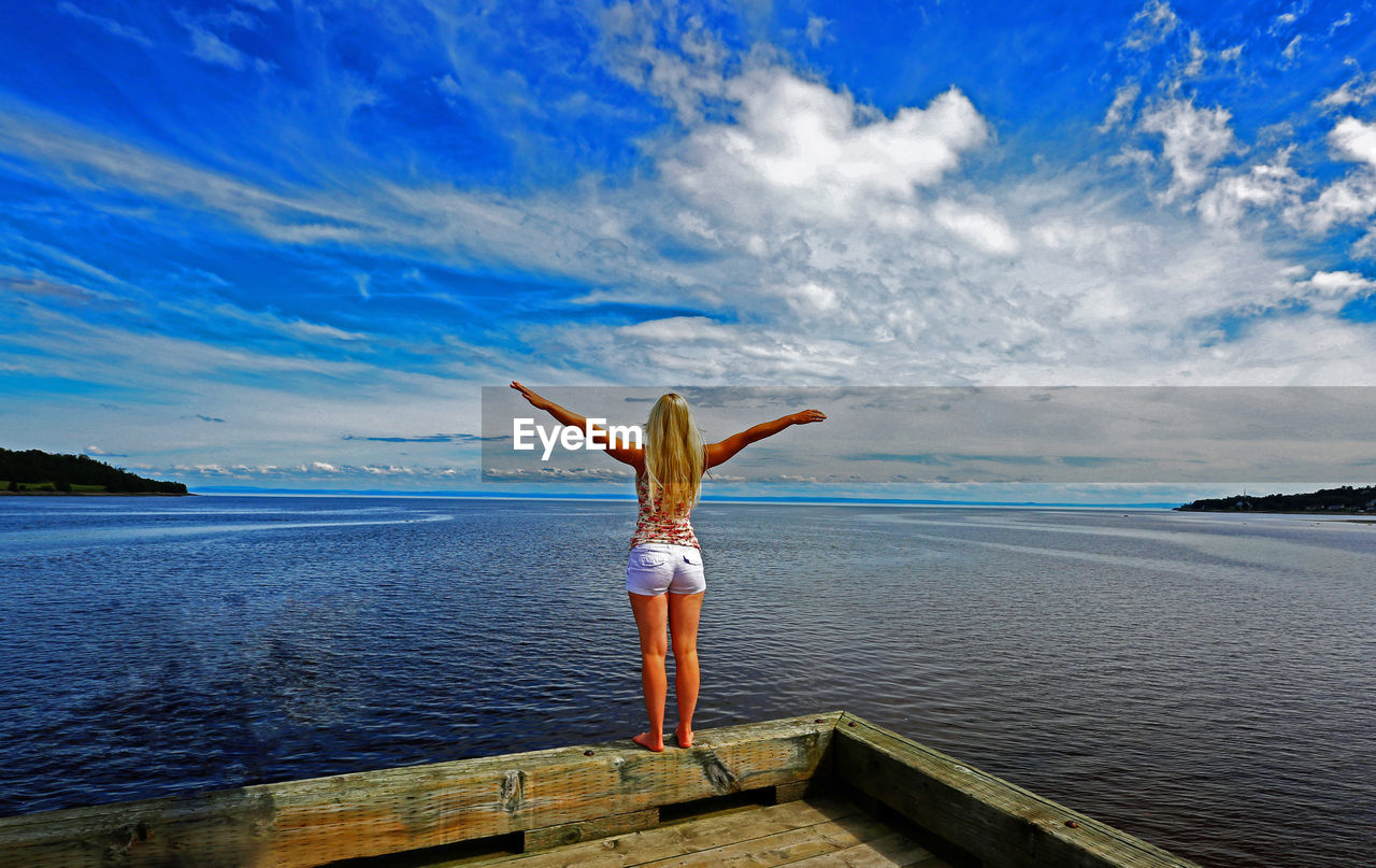 Rear view of young woman with arms outstretched standing on pier over sea against cloudy sky