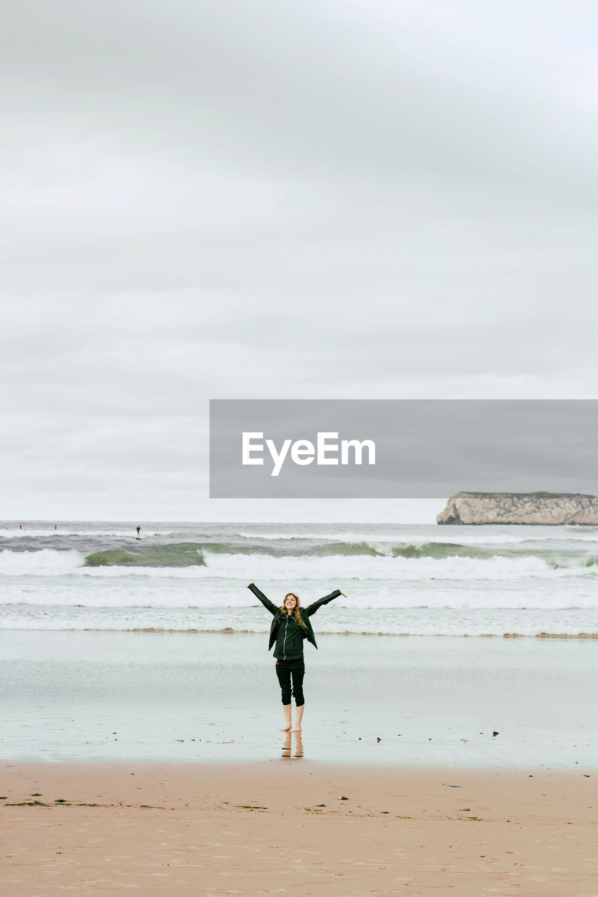 Full length of young woman with arms outstretched standing at beach against cloudy sky