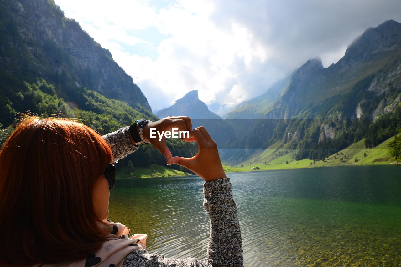 Rear view of woman making heart shape with hands over lake against mountains and sky