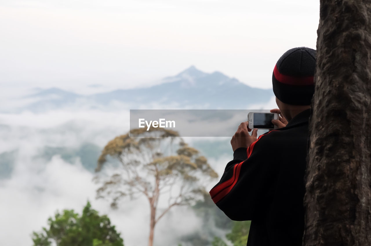 MIDSECTION OF PERSON PHOTOGRAPHING ON MOUNTAIN AGAINST SKY