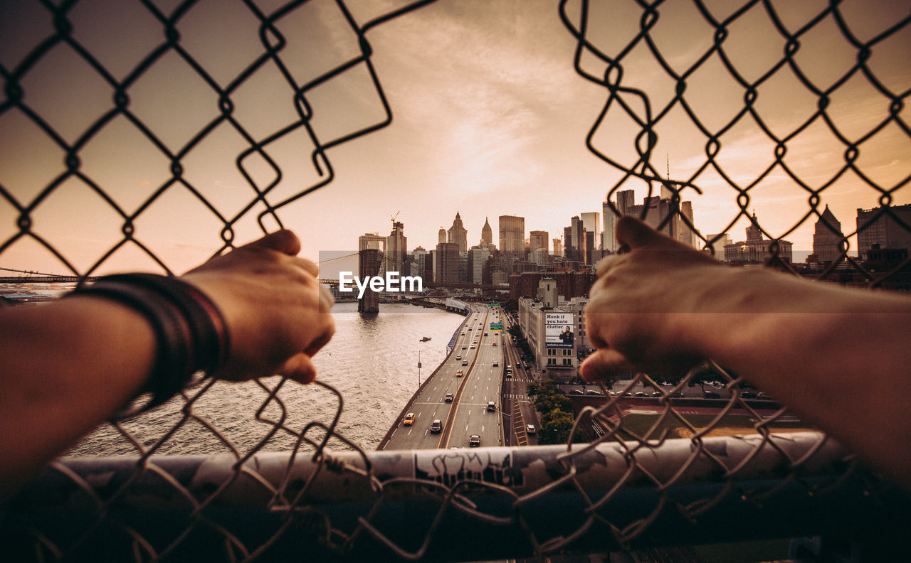 Cropped image of hands holding fence against sky in city