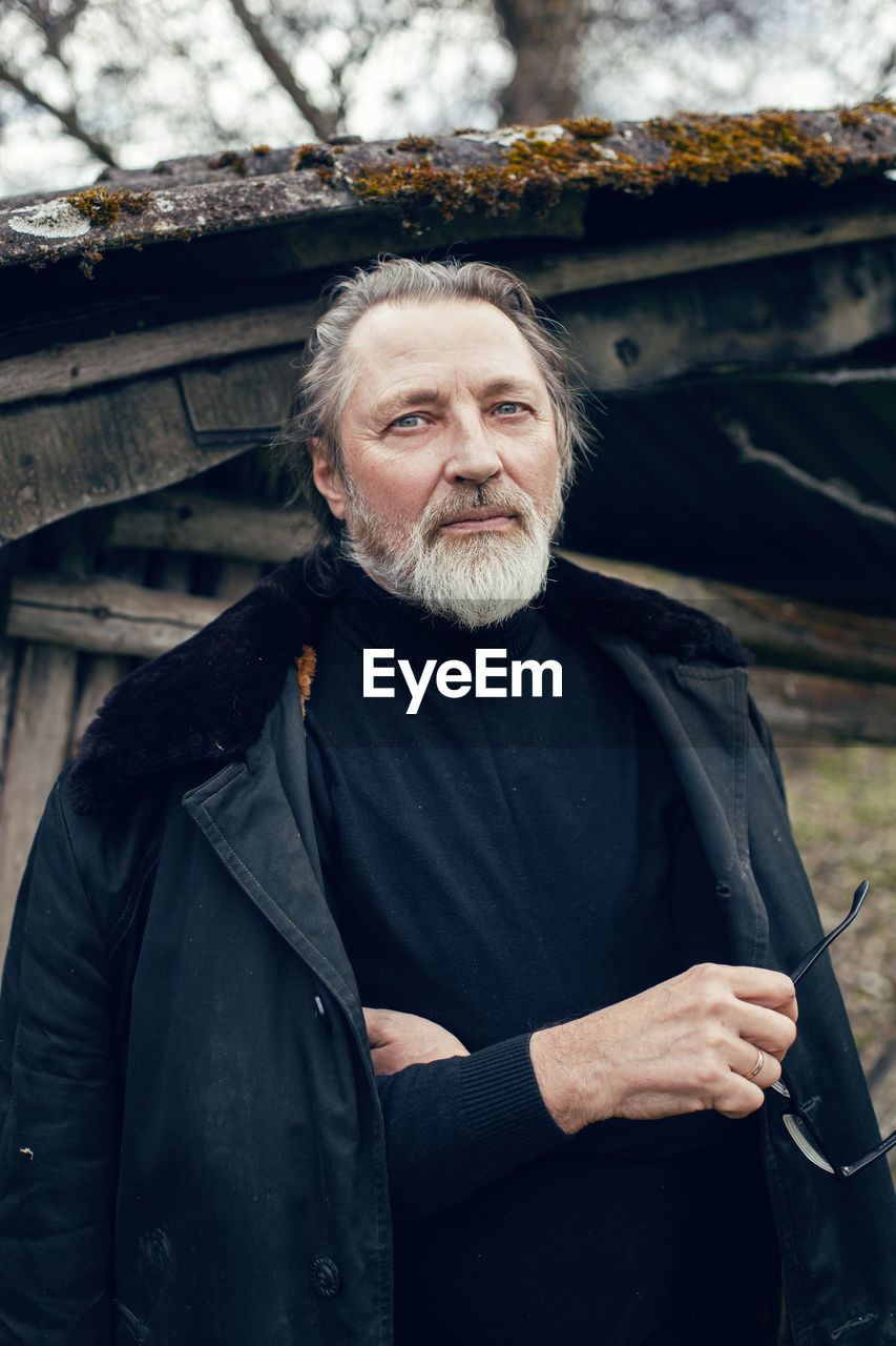 Elderly man with a beard stands in the village near a wooden shed in a sheepskin coat and glasses