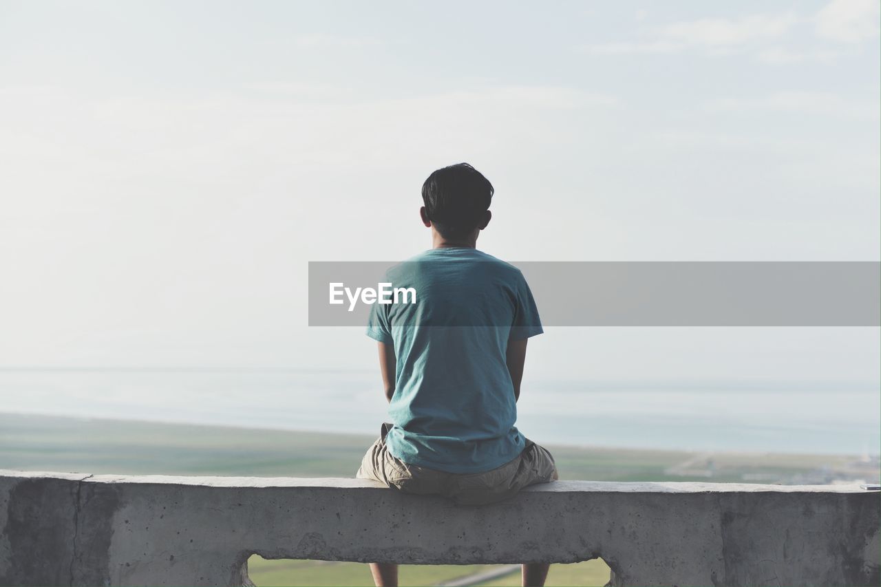 Rear view of man sitting on retaining wall against sky