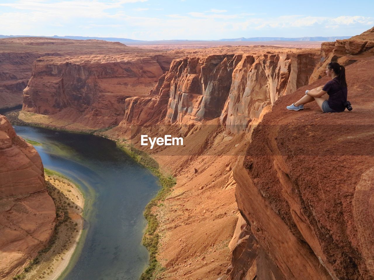 Full length of woman sitting on cliff by horseshoe bend against sky