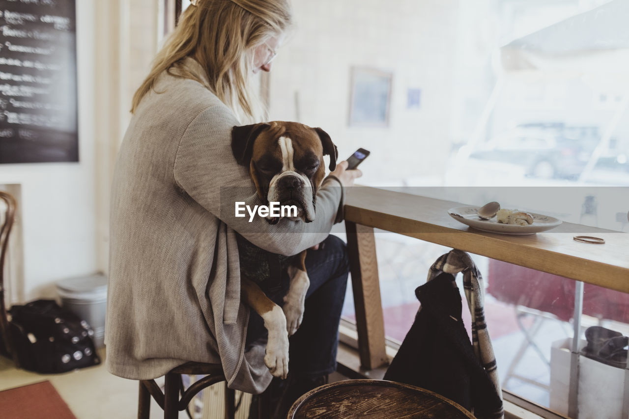 Side view of young woman using smart phone while sitting with dog at cafe