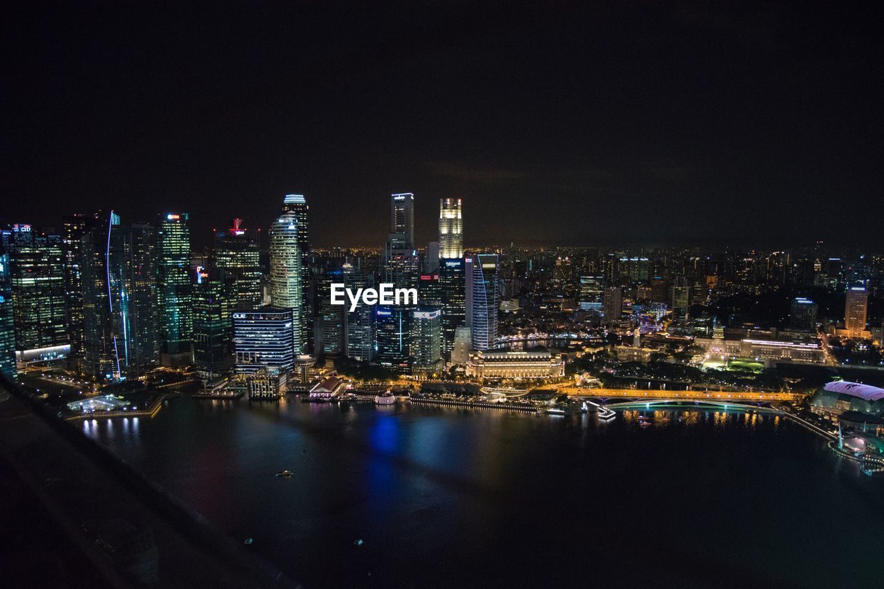 Illuminated buildings by river against sky at night