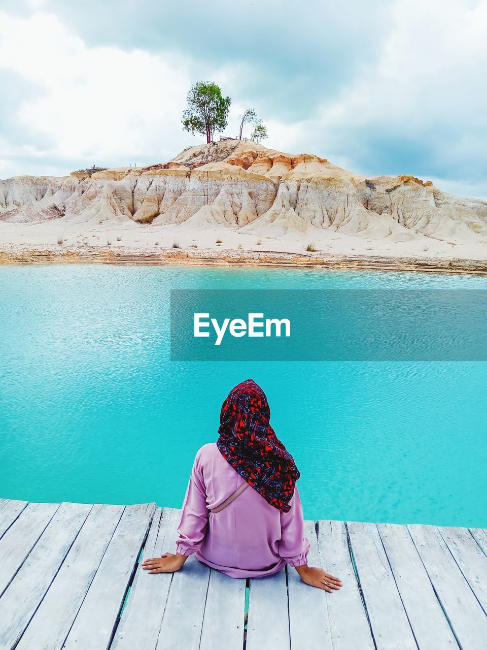 REAR VIEW OF WOMAN SITTING ON ROCK AGAINST SKY