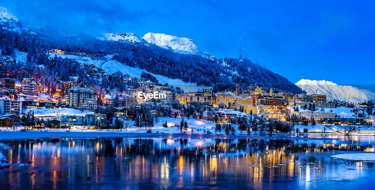 Buildings on snow covered mountain by lake against sky