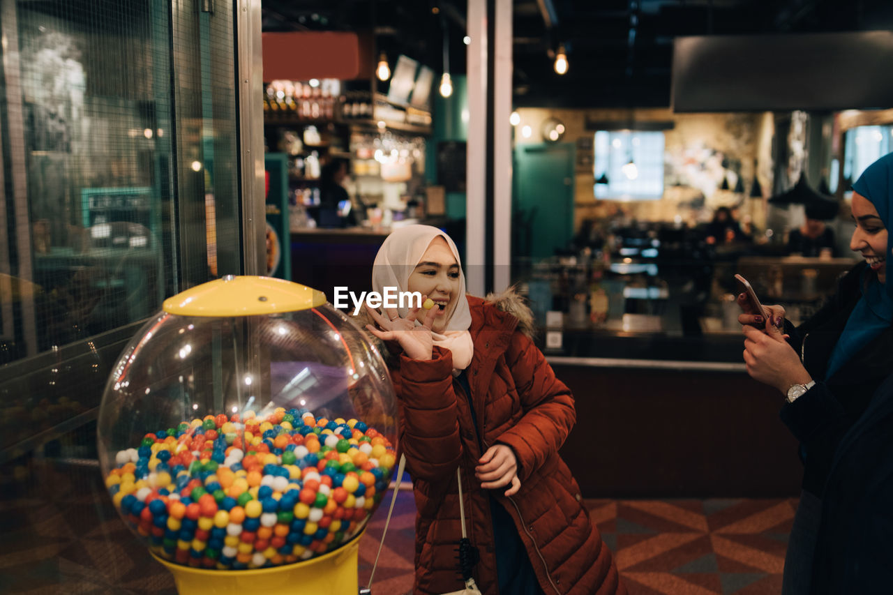 Young woman photographing cheerful female friend standing with bubble gum by vending machine