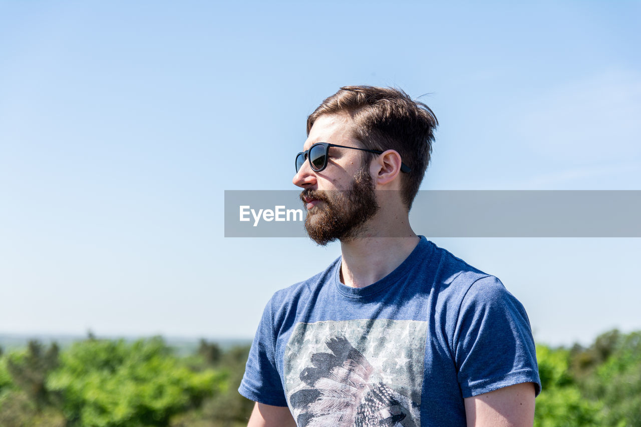 Bearded young man looking away while standing against sky during sunny day