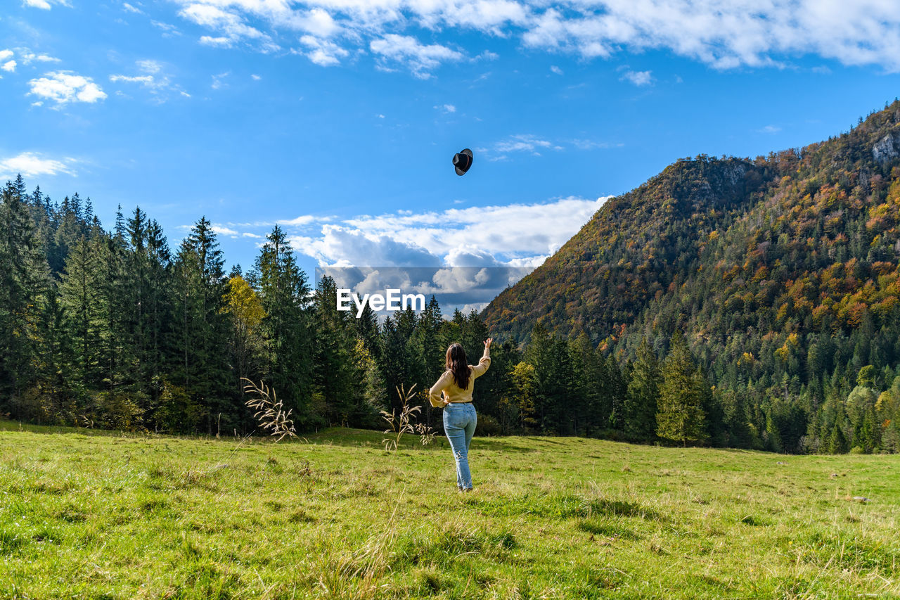 Rear view of young woman stading on green pasture, throwing hat in air.