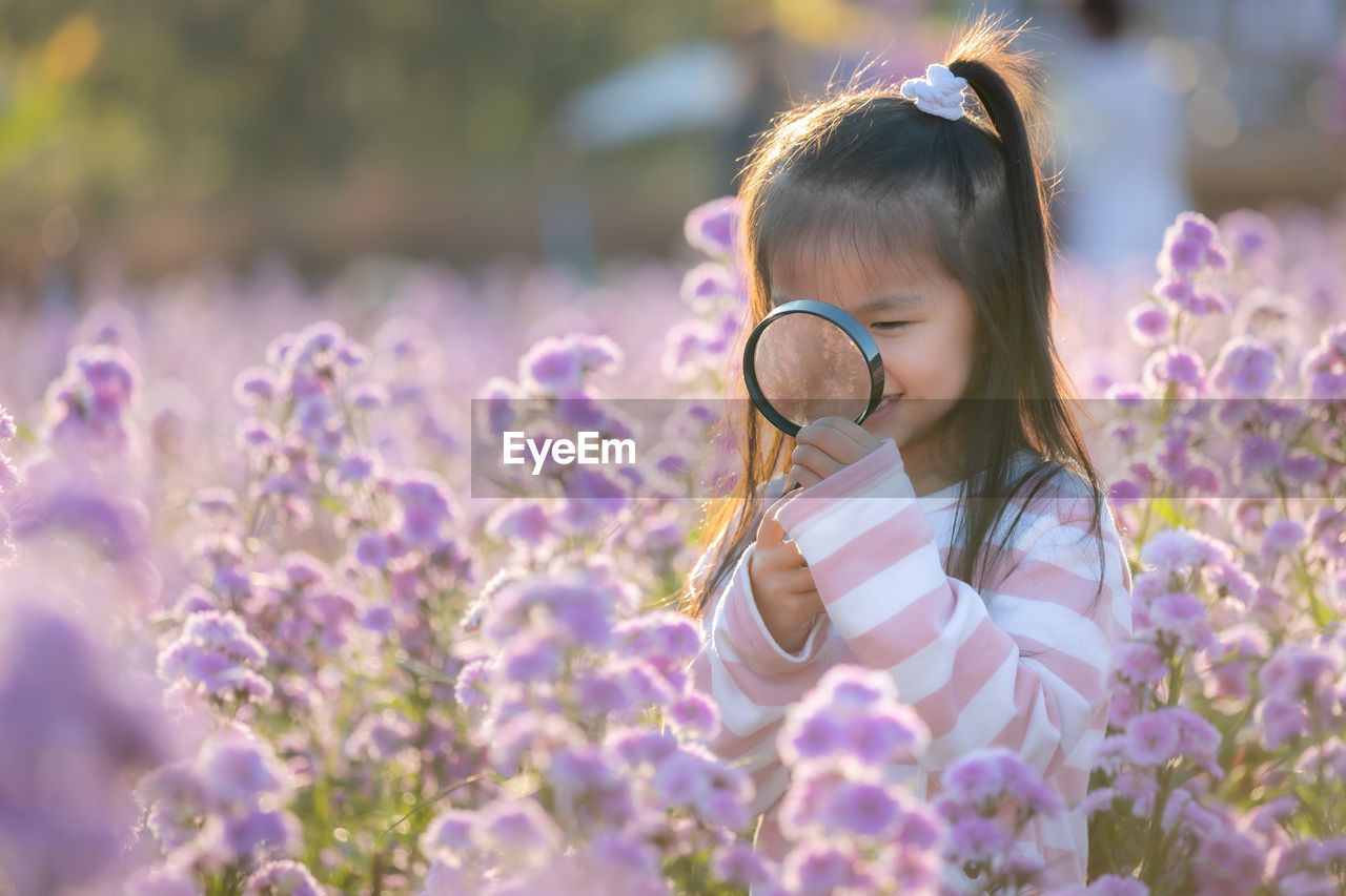 Close-up of girl holding magnifying glass by purple flowering plants