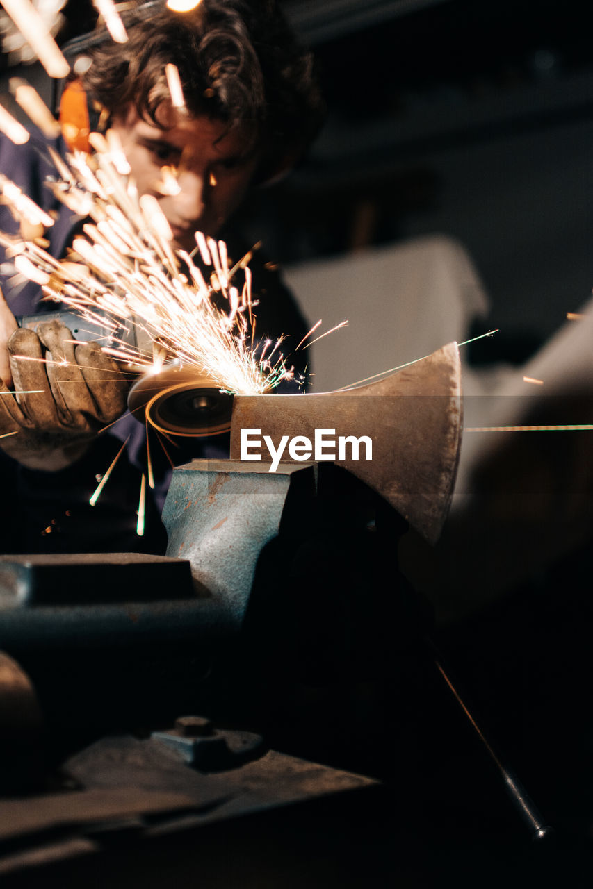 Focused male in gloves and headphones sharpening axe with electric grinder in flying sparkles in workshop