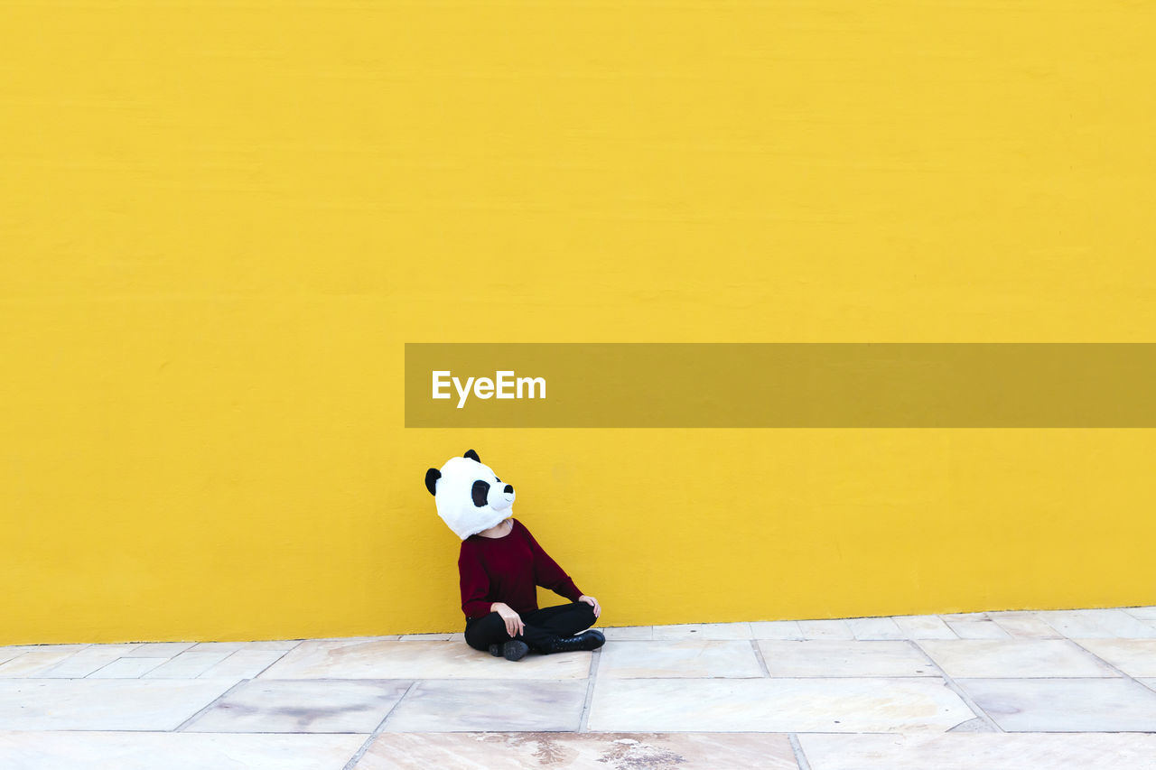 Woman wearing panda mask while sitting in cross-legged against yellow wall