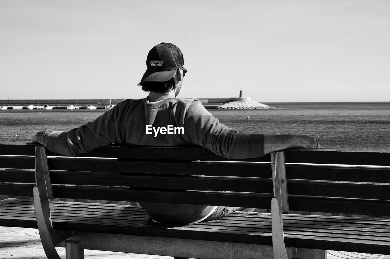 MAN SITTING ON BENCH BY SEA AGAINST SKY