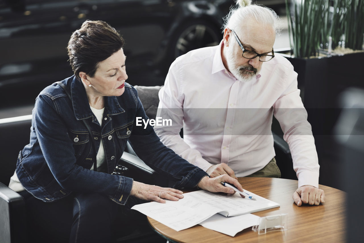 Senior man and woman analyzing documents while sitting at car showroom