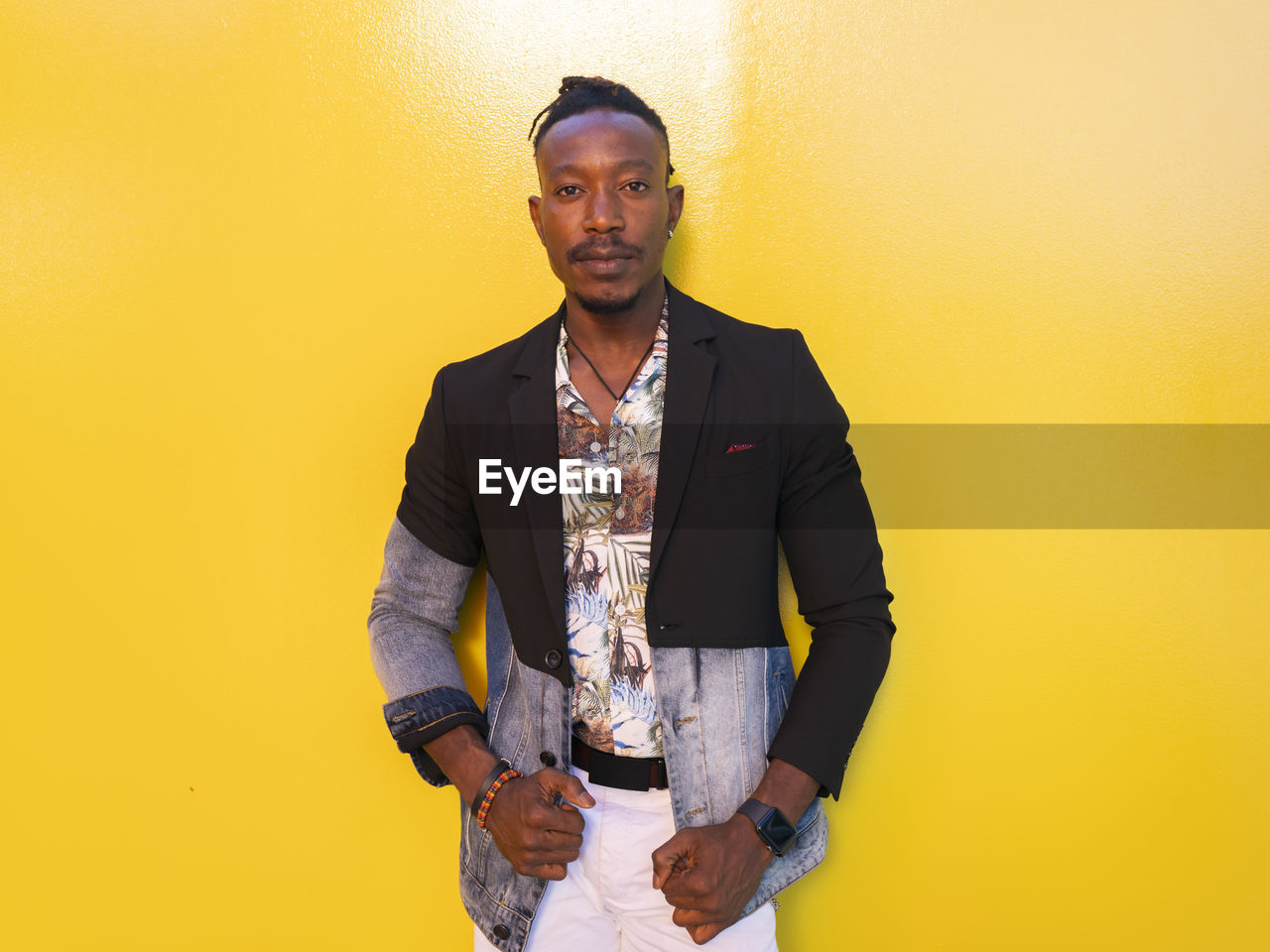 Serious african american male model in fancy outfit standing near yellow wall on street and looking at camera
