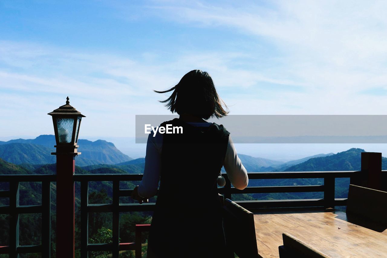 Rear view of woman standing by railing against mountains and sky