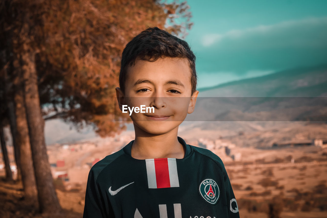 PORTRAIT OF SMILING BOY STANDING AGAINST BLURRED BACKGROUND