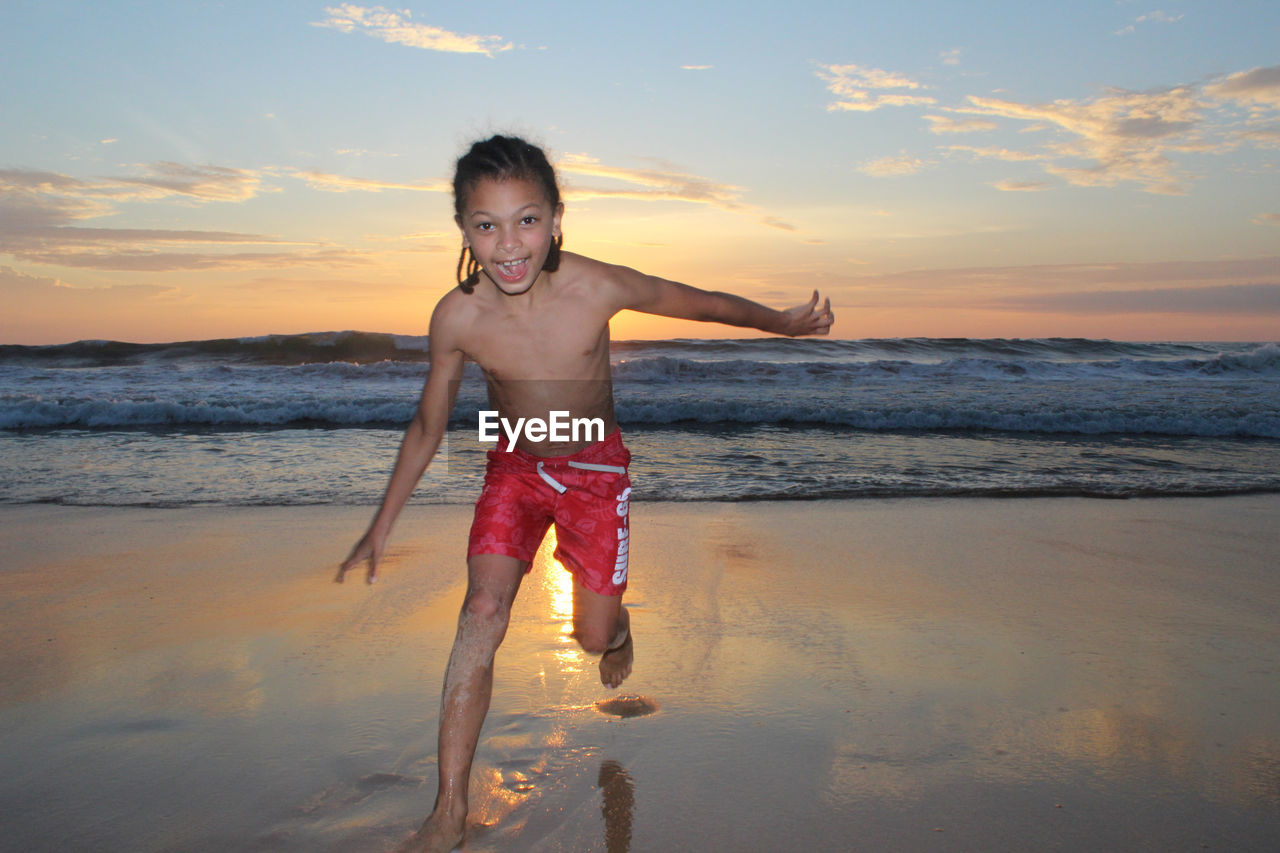 Portrait of smiling shirtless boy running on sea shore against sky during sunset