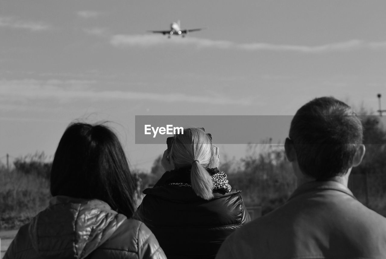 Rear view of people watching flying airplane against sky