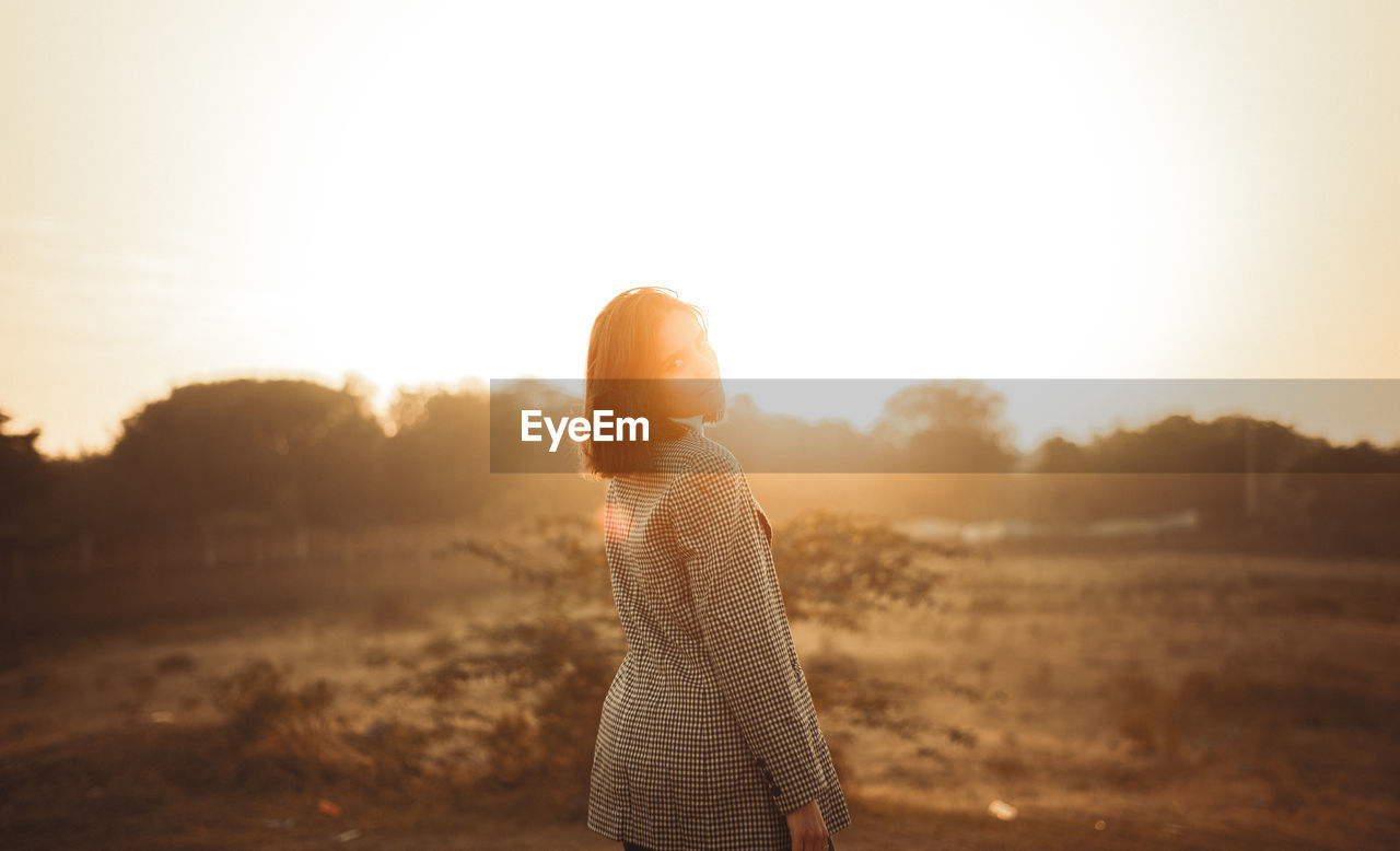 Side view of woman holding flower against sky during sunset