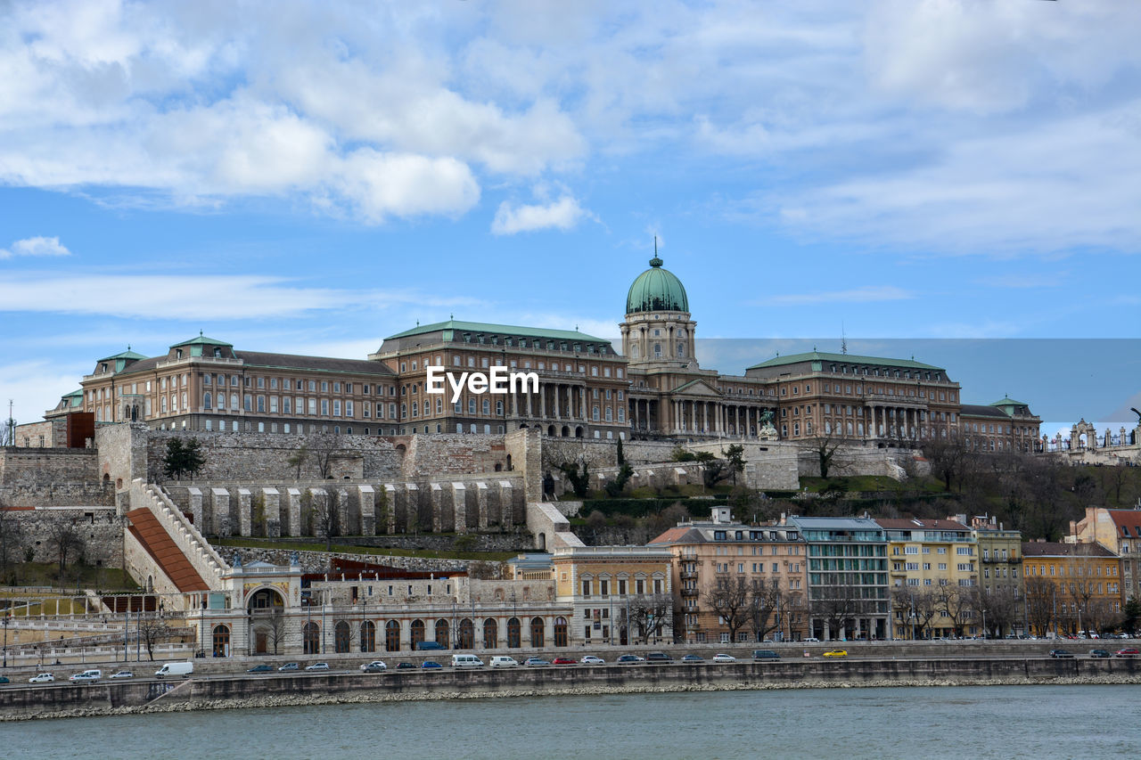 Danube river by buildings and buda castle against cloudy sky