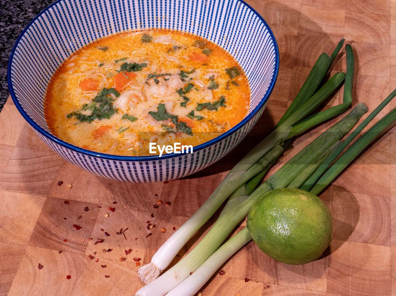 Asian coconut soup with red curry paste in a blue bowl on a wooden block with spring onions, lime.