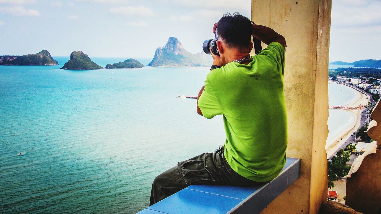 Rear view of man photographing mountains and sea while sitting on retaining wall