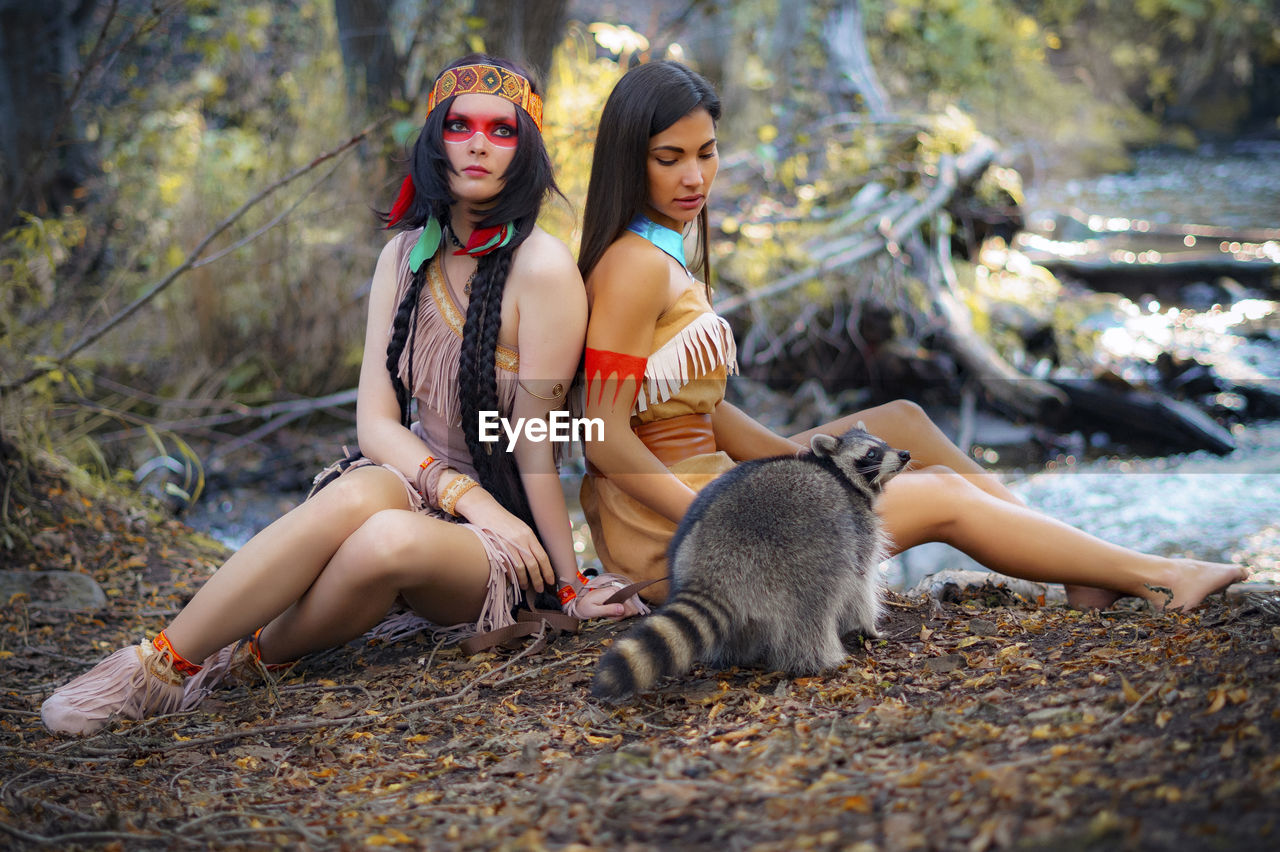 Young women in traditional clothing sitting by raccoon in forest