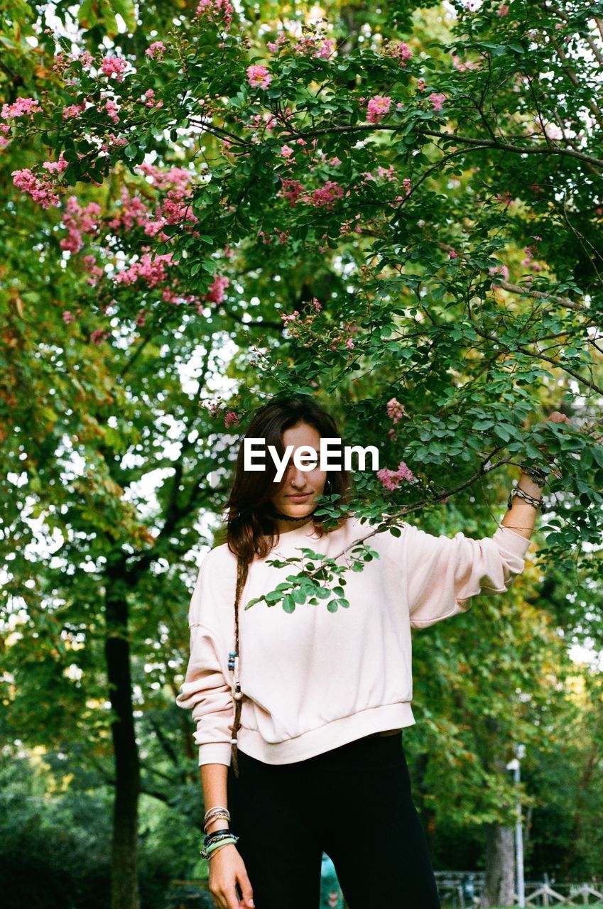 Portrait of young woman standing by trees at public park