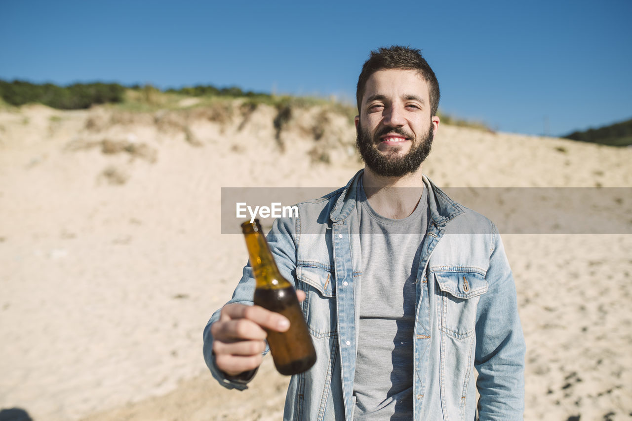 Young man holding alcoholic drink bottle while standing at beach during sunny day