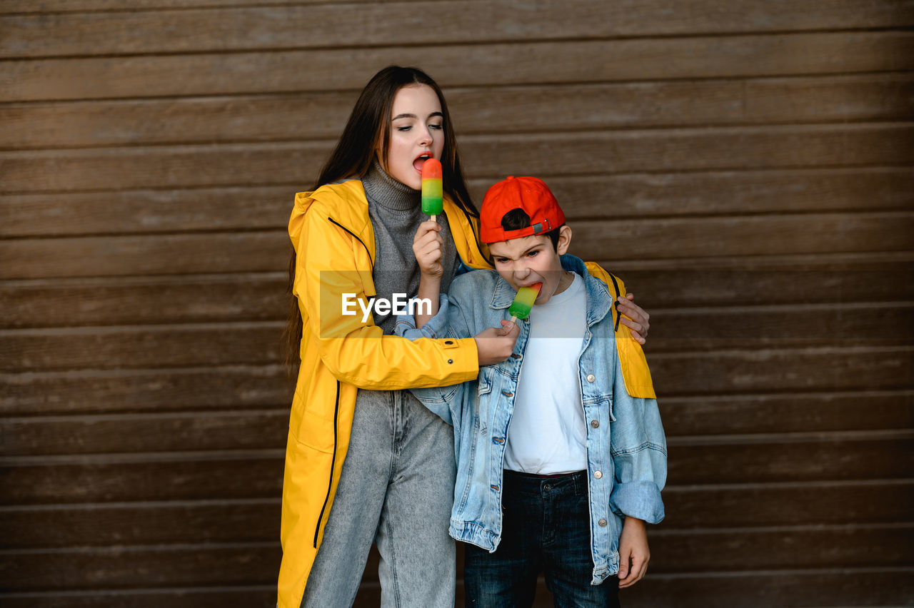 A girl in a yellow raincoat and a boy in a red cap are holding colorful ice cream in their hands