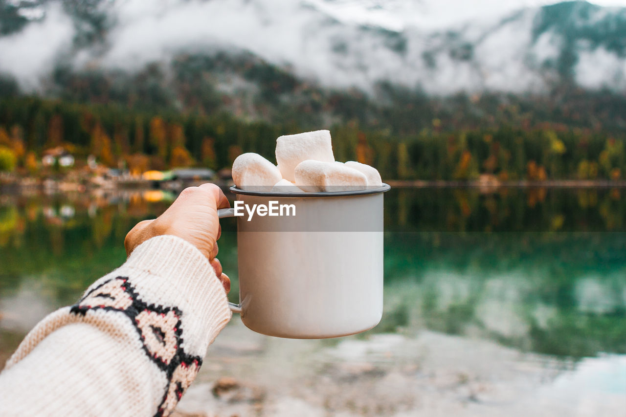 Picnic in bavarian mountains, germany. hand with mug of cacao and marshmallow on the lake background