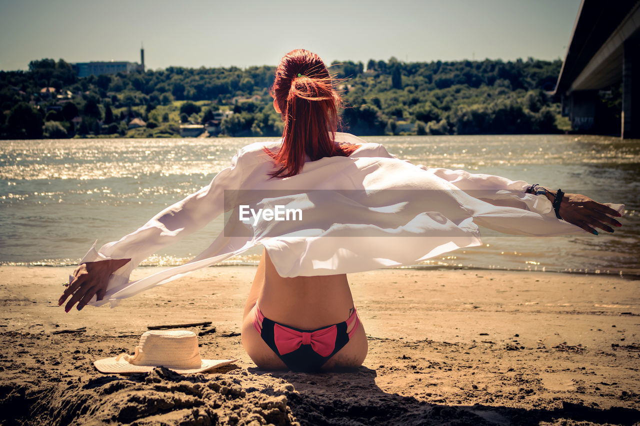 REAR VIEW OF WOMAN SITTING ON BEACH DURING SUNNY DAY