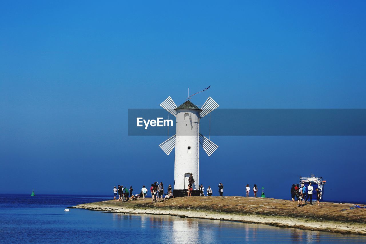 PEOPLE ON LIGHTHOUSE BY SEA AGAINST CLEAR SKY