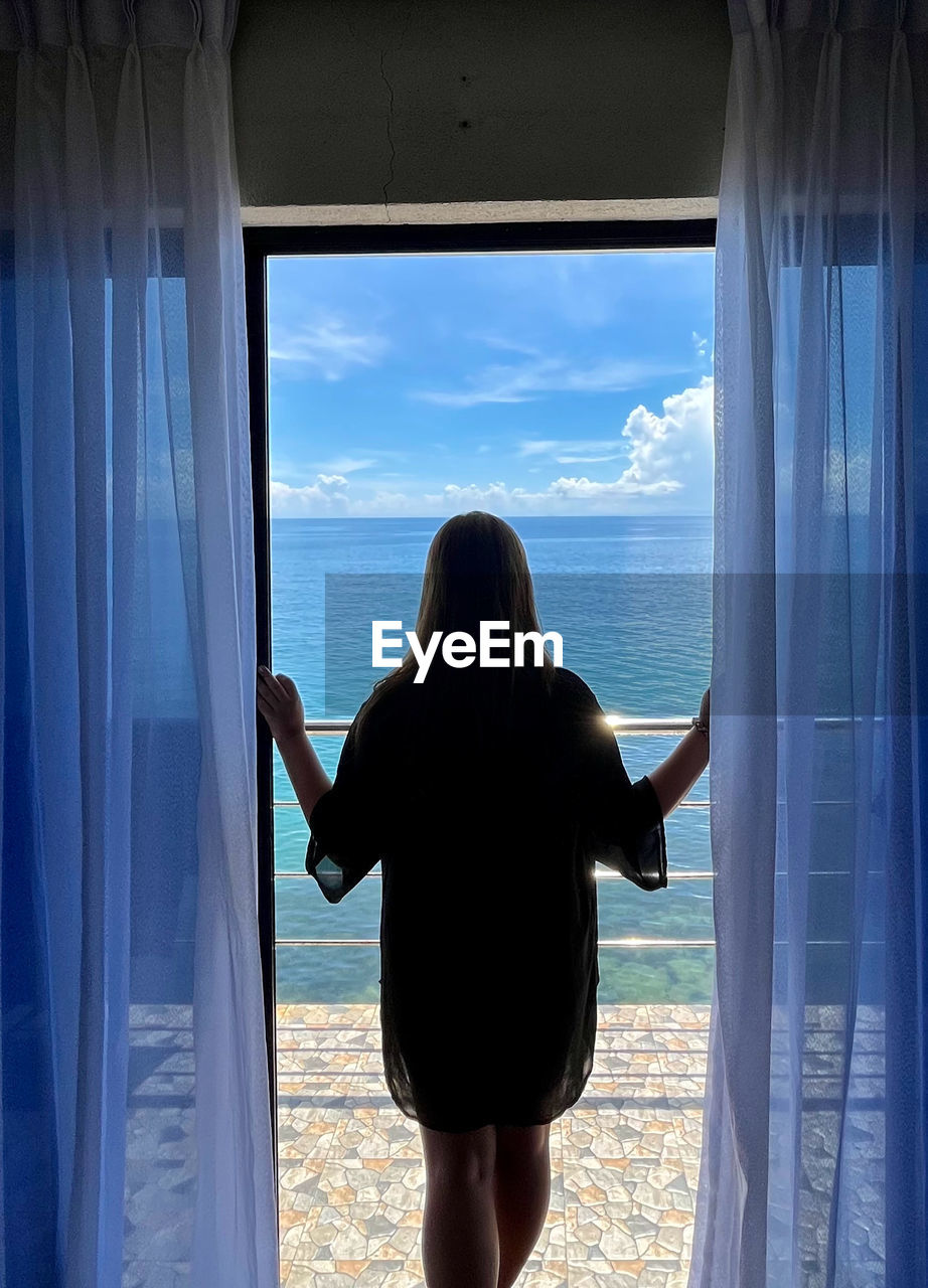 blue, rear view, one person, window, sea, adult, women, curtain, vacation, water, holiday, trip, standing, nature, looking through window, looking at view, sky, leisure activity, lifestyles, looking, indoors, day, travel, beach, land, horizon, beauty in nature, scenics - nature, silhouette, tranquility, young adult, solitude, dress, three quarter length, horizon over water, long hair, relaxation, sunlight, hairstyle, interior design, window treatment, tranquil scene, female, glass