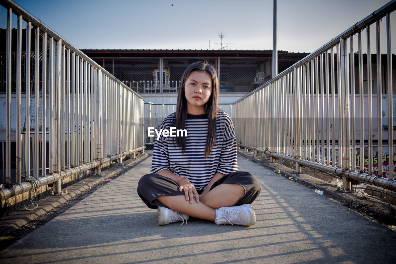 Full length portrait of teenage girl sitting on footbridge