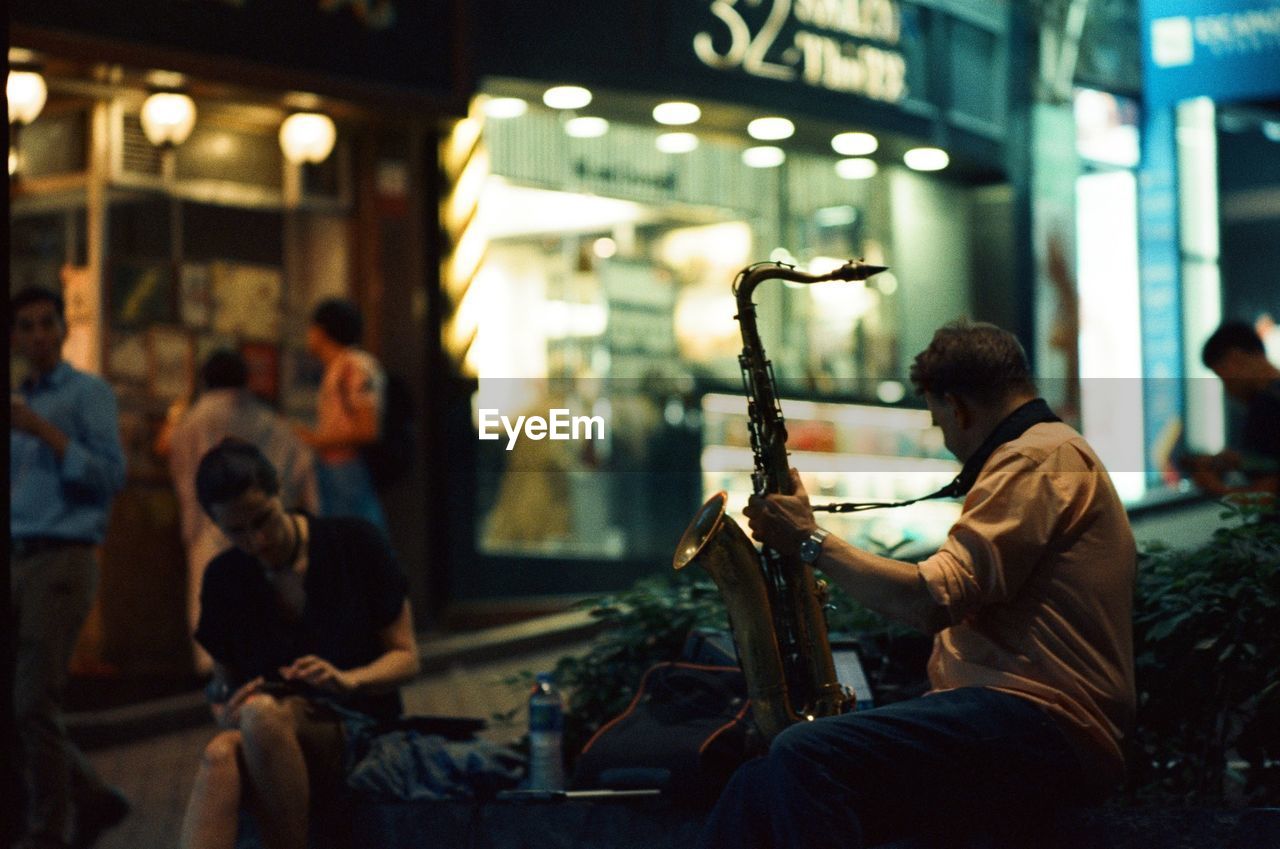 Side view of man holding saxophone while sitting in illuminated city at night