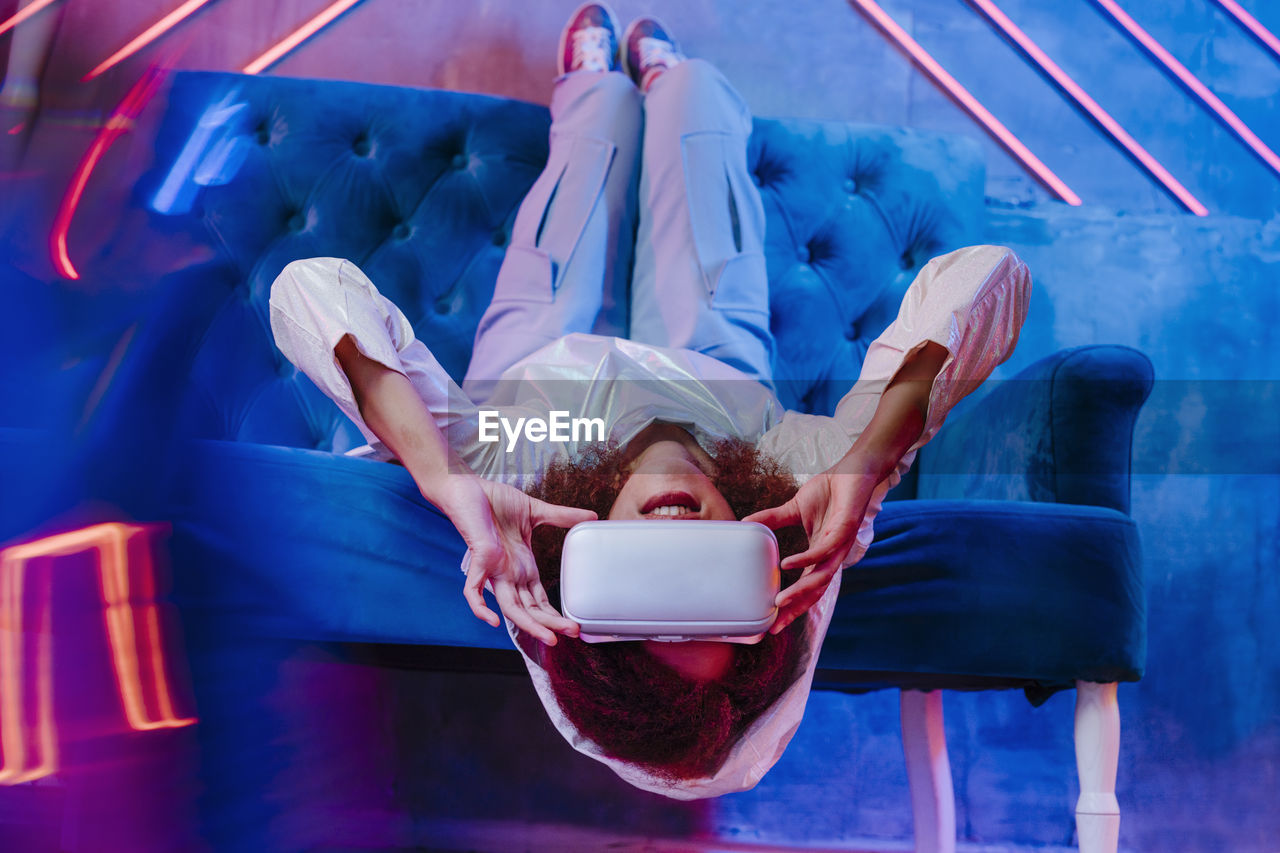 Woman with vr glasses lying upside down on sofa in front of neon light