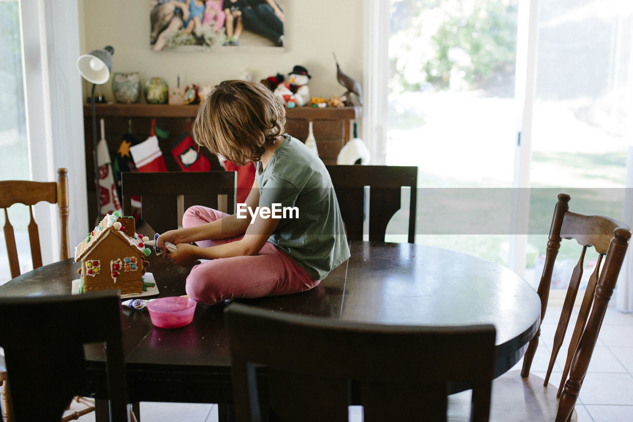 Girl making gingerbread house on table at home