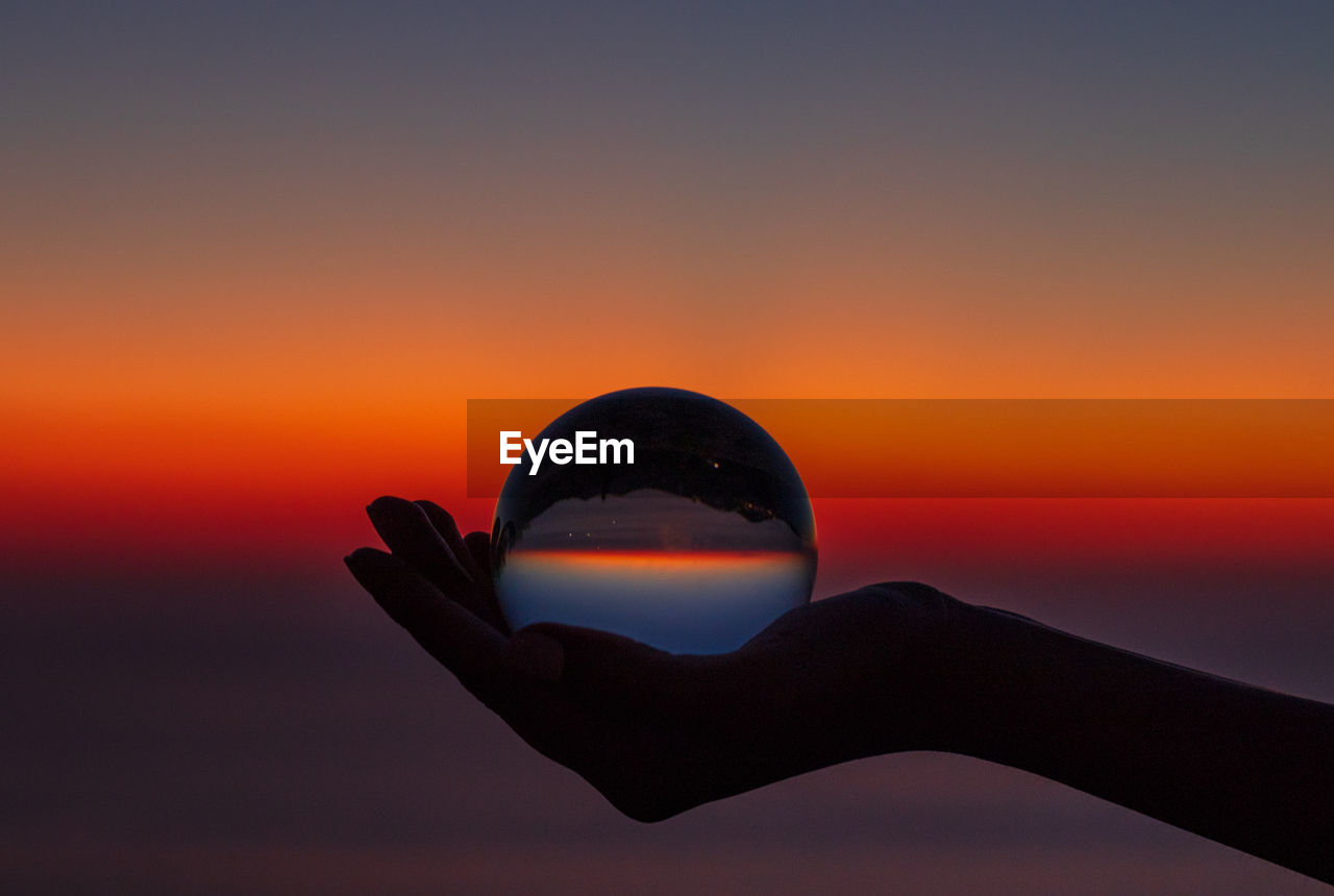 Cropped hand holding crystal ball against sky during sunset