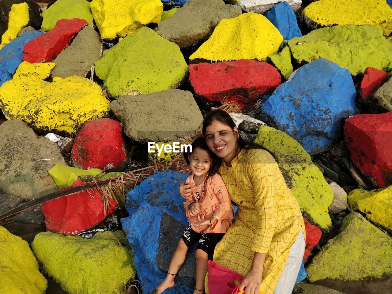 Portrait of a smiling young woman with daughter sitting on colorful rocks at beach